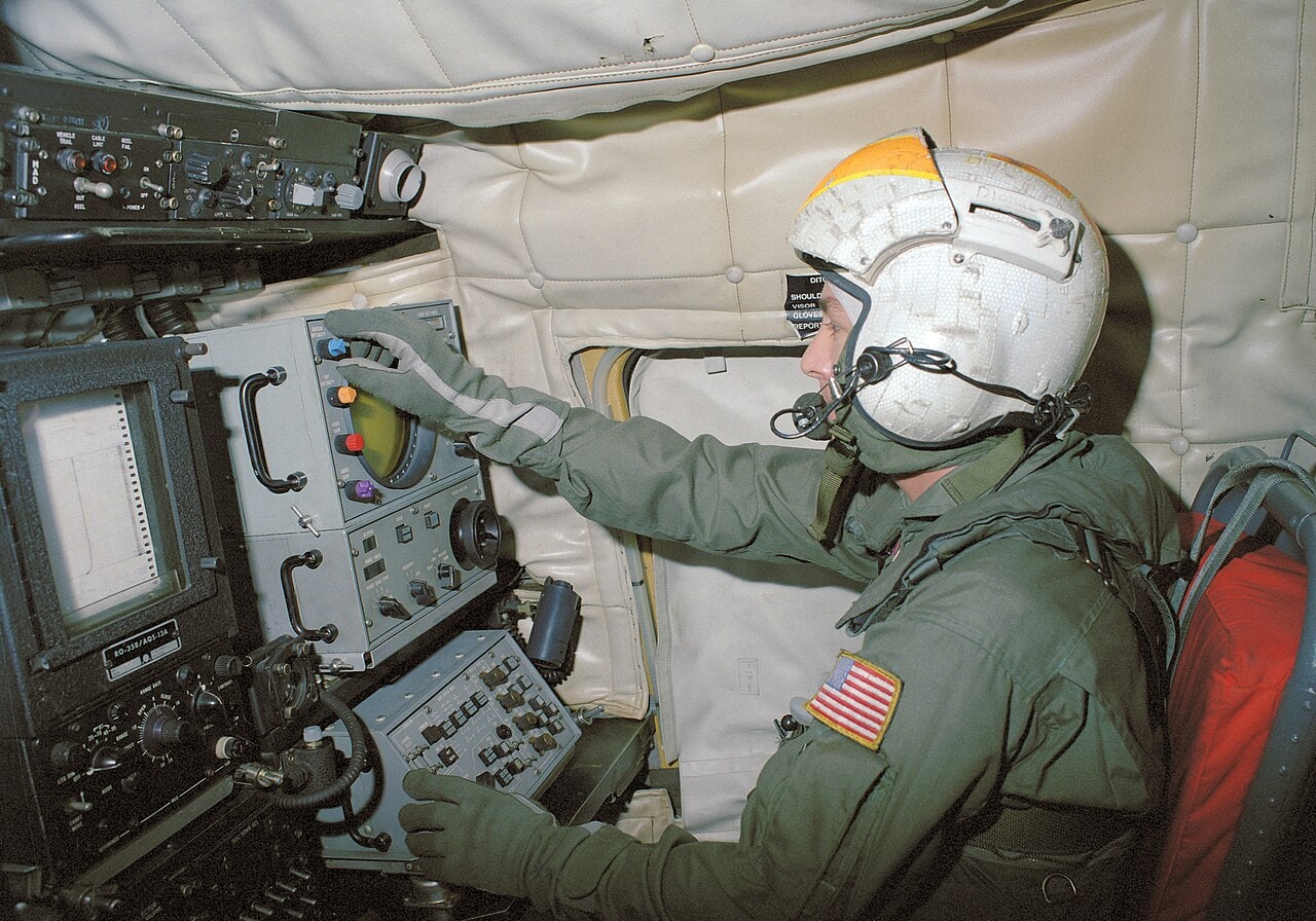 Militar da Marinha dos EUA usando capacete branco e macacão verde opera painel de sonar com radar circular dentro de helicóptero Sea King em 1987.