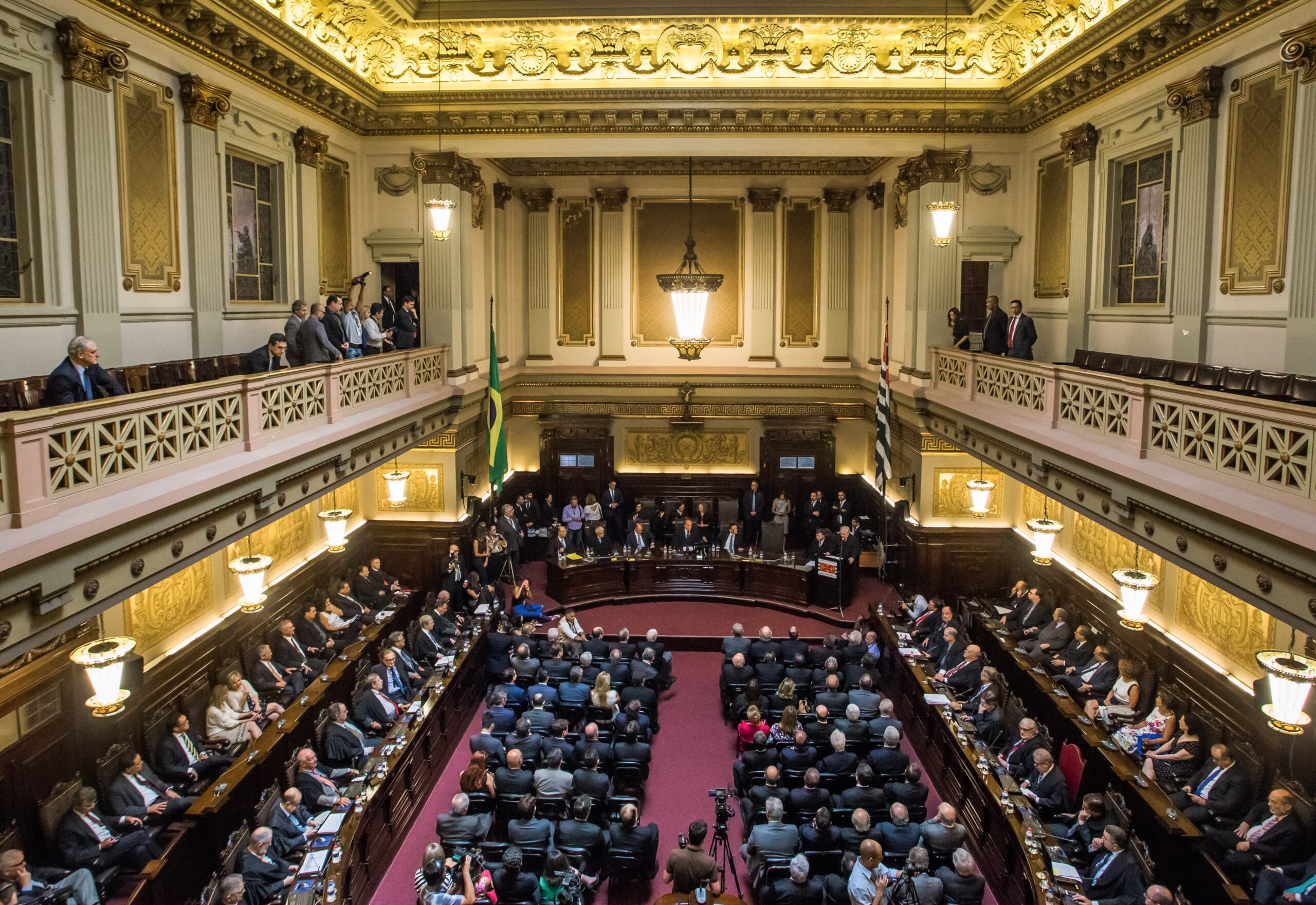 Sessão solene no salão do Palácio da Justiça de São Paulo, com galerias, lustre e dezenas de pessoas em cadeiras.
